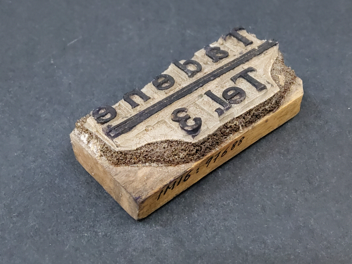 Stämpel, rektangulär av trä med gummibokstäver: "Tådene tel. 3".