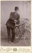 Porträtt av en okänd man med hatt som står lutad mot en cykel.