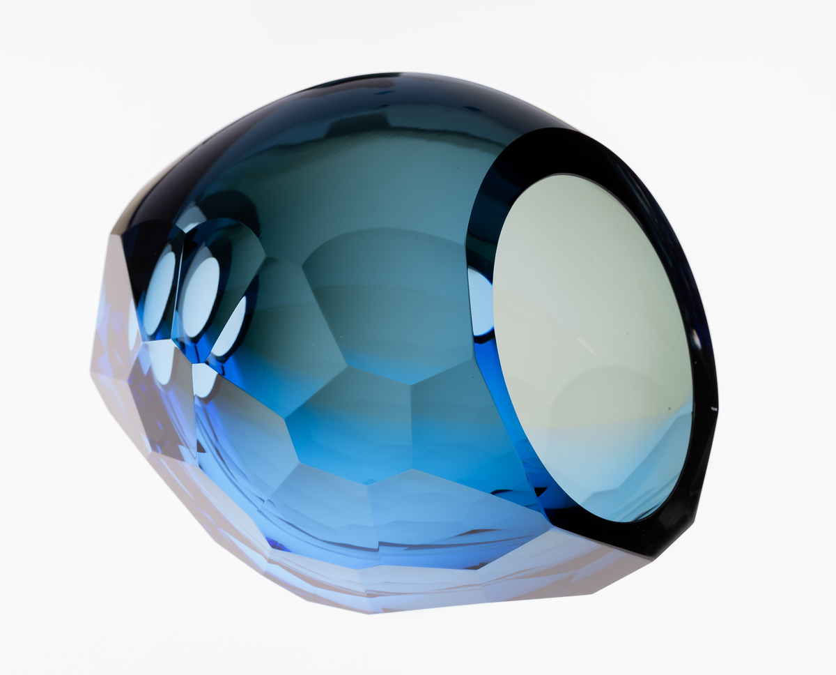 Ovalformet skulptur i farget gjennomskinnelig glass. Yttersiden er fasettert ved hjelp av sliping og har sirkulære åpninger på hver av kortsidene. Massen graderes fra grå mot blå, og overflaten veksler mellom mørke og lysere partier.