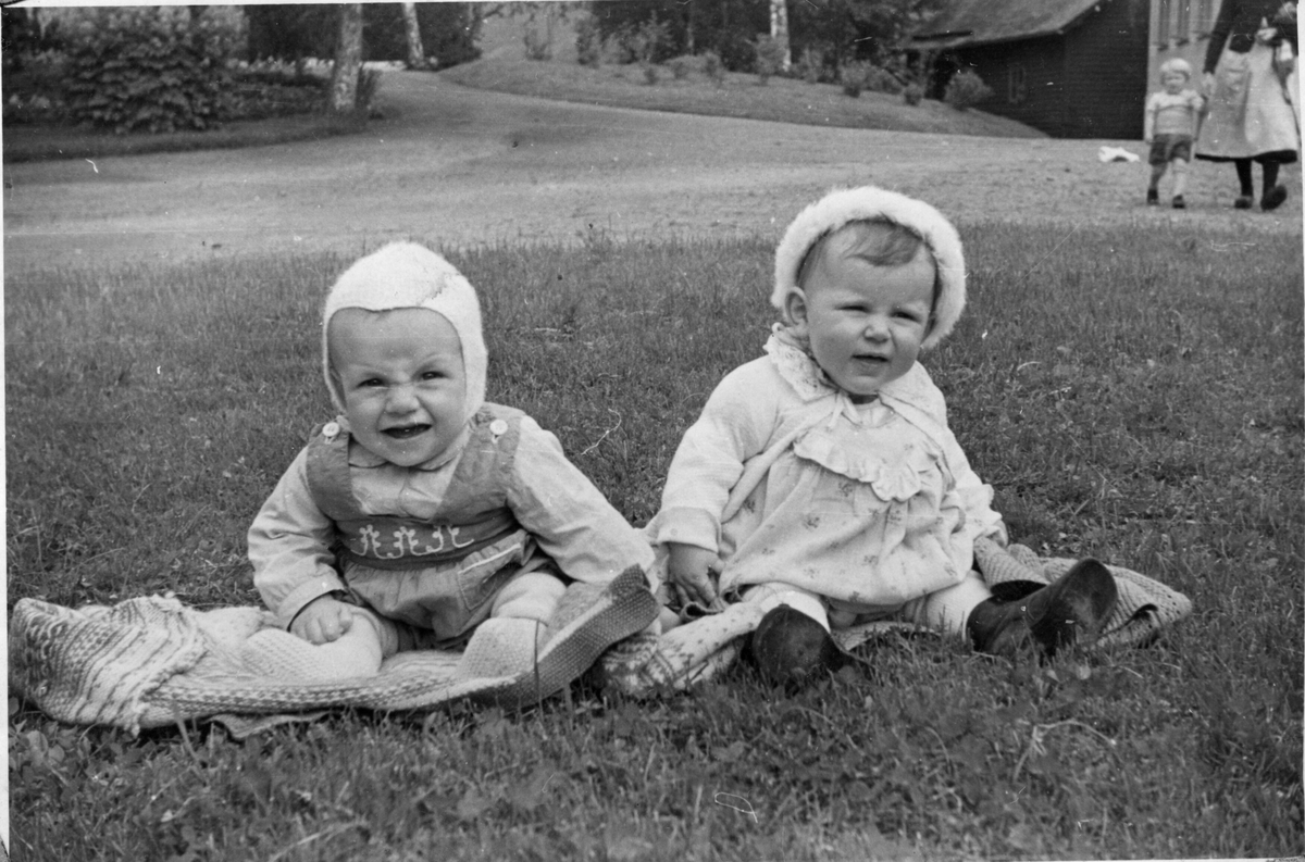 To små born som sit på graset. Ei kvinne med eit born  i handa i bakgrunnen.