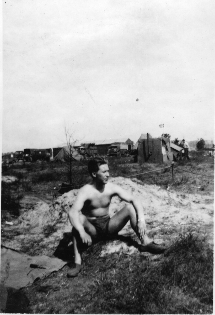 Ein ung mann i kortbukse og sko som sit på bakken.  Militærekøyretøy i bakgrunnen. Fotografert i tilknyting til invasjonen i Normandie.