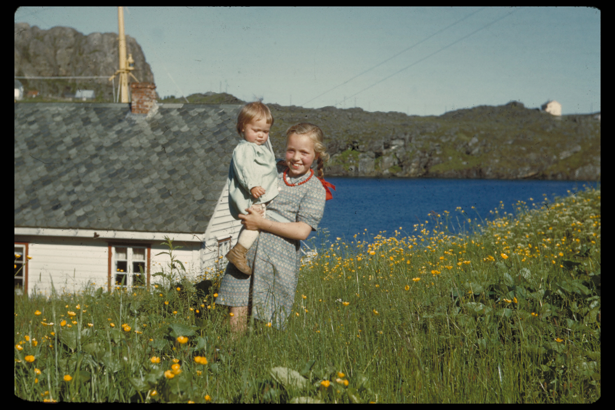 Telavåg etter gjenreisinga. Ei jente med ei lita jente på armen i ei blomstereng på Øvretveit Eit naust og Nipafjellet i bakgrunnen.