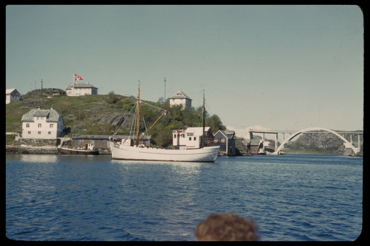 Telavåg etter gjenreisinga. Bustadhus og naust på Telle. To fiskeskøyter ligg til kai og på reia utanfor. Brua over Telasundet i bakgrunnen. Det norske flagget er heist ved eit bustadhus.
