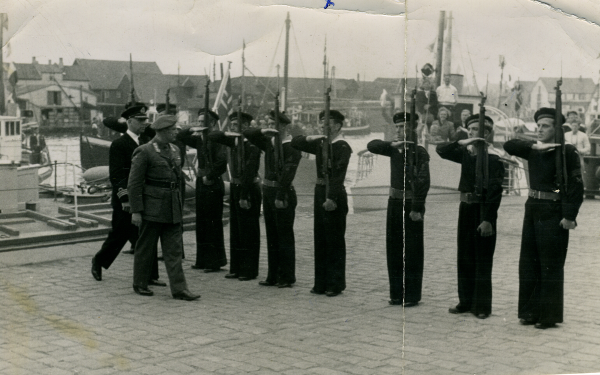 Ni marinegastar som står æresvakt for Kronprins Olav  og ein marineoffiser på ein kai i Haugesund.