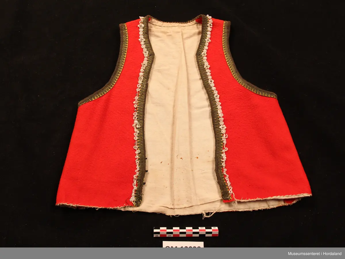 Raudt bunadsliv med gule og grøne silkekvardar. Innom kvarden, framme og rundt hals, er det påsydde perlesnorer der perlene dannar blomstermønster. Tett halsskjering. Tre hekter til attlat nede. Ubleika bomullsfor. Overkasta sårkant nede. Livet er sydd inn under armane.