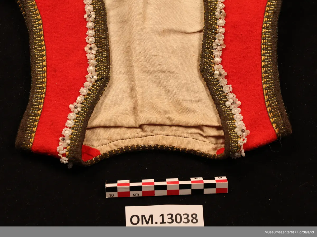 Raudt bunadsliv med gule og grøne silkekvardar. Innom kvarden, framme og rundt hals, er det påsydde perlesnorer der perlene dannar blomstermønster. Tett halsskjering. Tre hekter til attlat nede. Ubleika bomullsfor. Overkasta sårkant nede. Livet er sydd inn under armane.