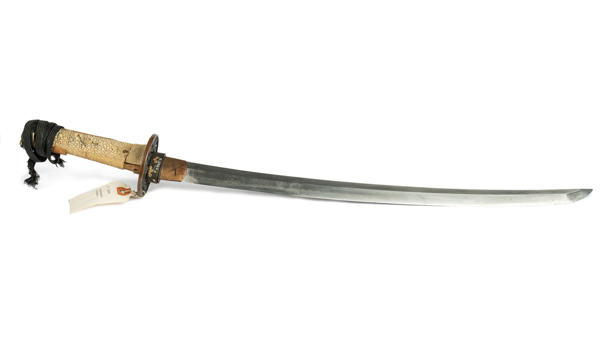 Samurajsvärd från Japan. Klinga svagt bakåtböjd, eneggad skarp, facetterad rygg. Fäste av vitt hajskinn (stingrocka?), flätad med svart bomullsgarn samt försett med två amuletter, den ena med guldinläggningar. Fästets beslag upptill och nedtill har förgyllda inläggningar. Klinga märkt: Lindman. Parerplåt rund, genombrutet bladverk med fåglar.
