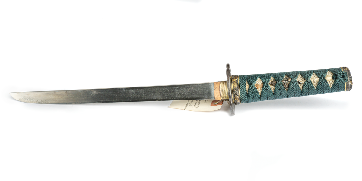 Samurai-dolk (WAKICACHI) från Japan. Klinga svagt bakåtböjd,eneggad, skarp, facetterad rygg. Fäste av vitt hajskinn flätat med grönt bomullsband med två amuletter, delvis förgyllda. Beslag upptill och nedtill cicilerade och förgyllda. Parerplåt rund med ett hål på ena sidan samt med förgyllt och cicilerat bladverk. Balja av trä, svartlackerad med beslag å fyra ställen.
Låsfjädern,d.v.s. bestickknivens handtag saknas.
Kniven på baljan (KOZUKA) saknas.
Från fregatten VANADIS världsomsegling 1883-85.