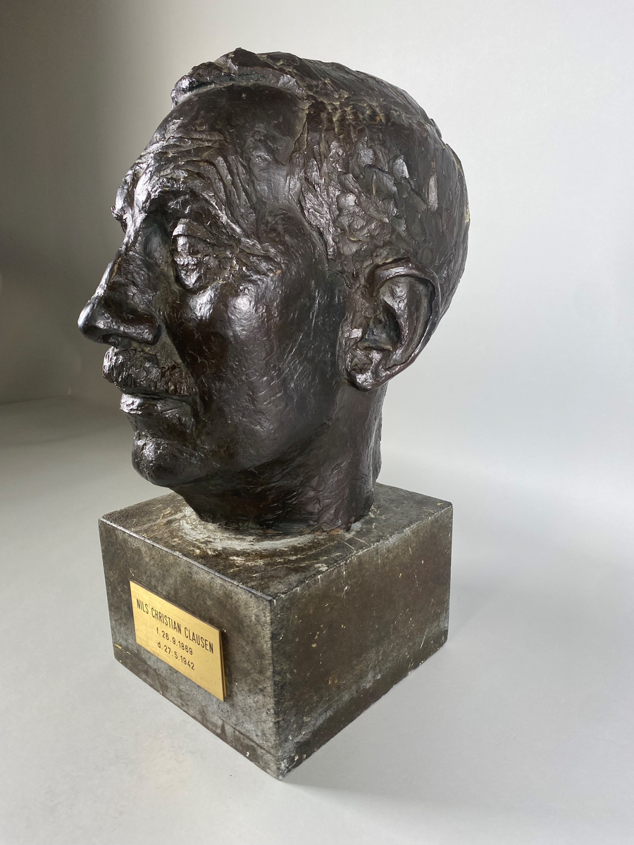 Bronsebyste av Nils Christian Clausen (1869-1942). Festet med en lang skrue til en sokkel av stein med navneplate.