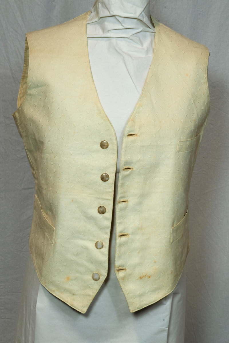 Offwhite enkeltspent vest uten krage, med tilhørende tversoversløyfe. Fronten er lin(?), eller aida bomullstoff med broderier i samme farge. Rygg og for er bomull. Metallknapper (tinn?). Spenne i ryggen. Brukt av brudgom i 1921.