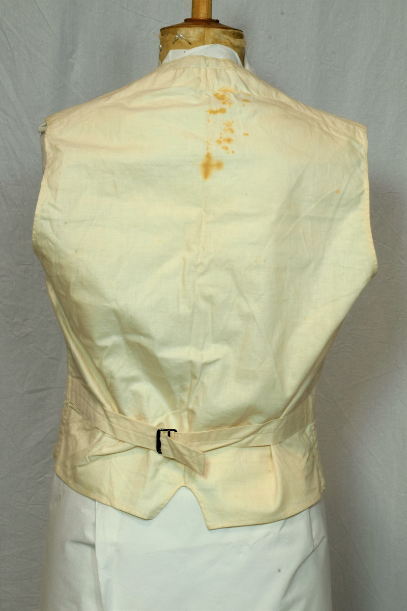 Offwhite enkeltspent vest uten krage, med tilhørende tversoversløyfe. Fronten er lin(?), eller aida bomullstoff med broderier i samme farge. Rygg og for er bomull. Metallknapper (tinn?). Spenne i ryggen. Brukt av brudgom i 1921.