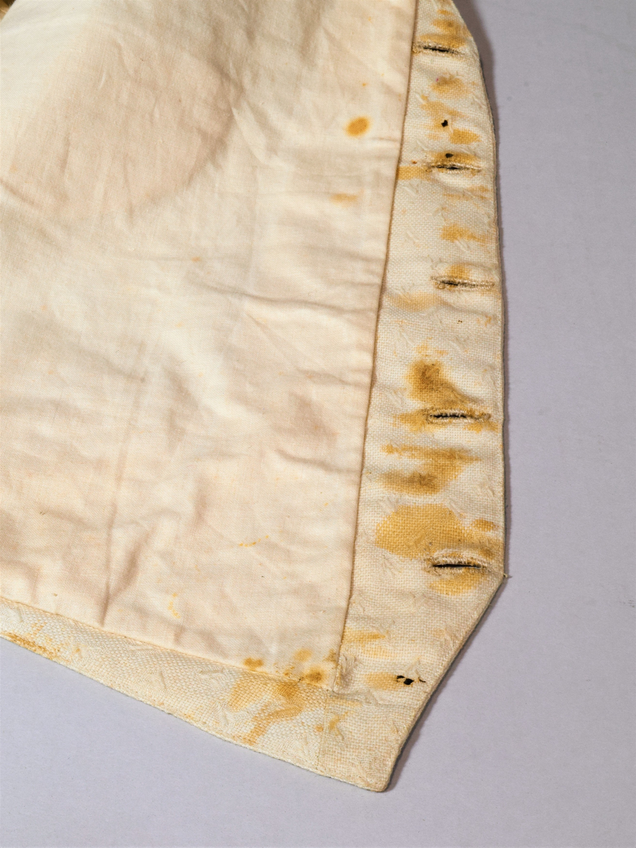 Offwhite enkeltspent vest uten krage, med tilhørende tversoversløyfe. Fronten er lin(?), eller aida bomullstoff med broderier i samme farge. Rygg og for er bomull. Metallknapper (tinn?). Spenne i ryggen. Brukt av brudgom i 1921.