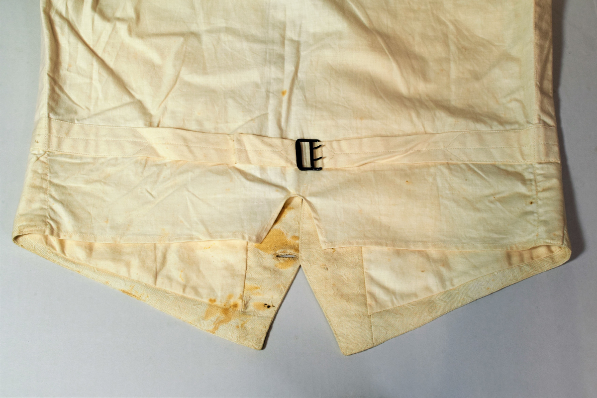 Offwhite enkeltspent vest uten krage, med tilhørende tversoversløyfe. Fronten er lin(?), eller aida bomullstoff med broderier i samme farge. Rygg og for er bomull. Metallknapper (tinn?). Spenne i ryggen. Brukt av brudgom i 1921.