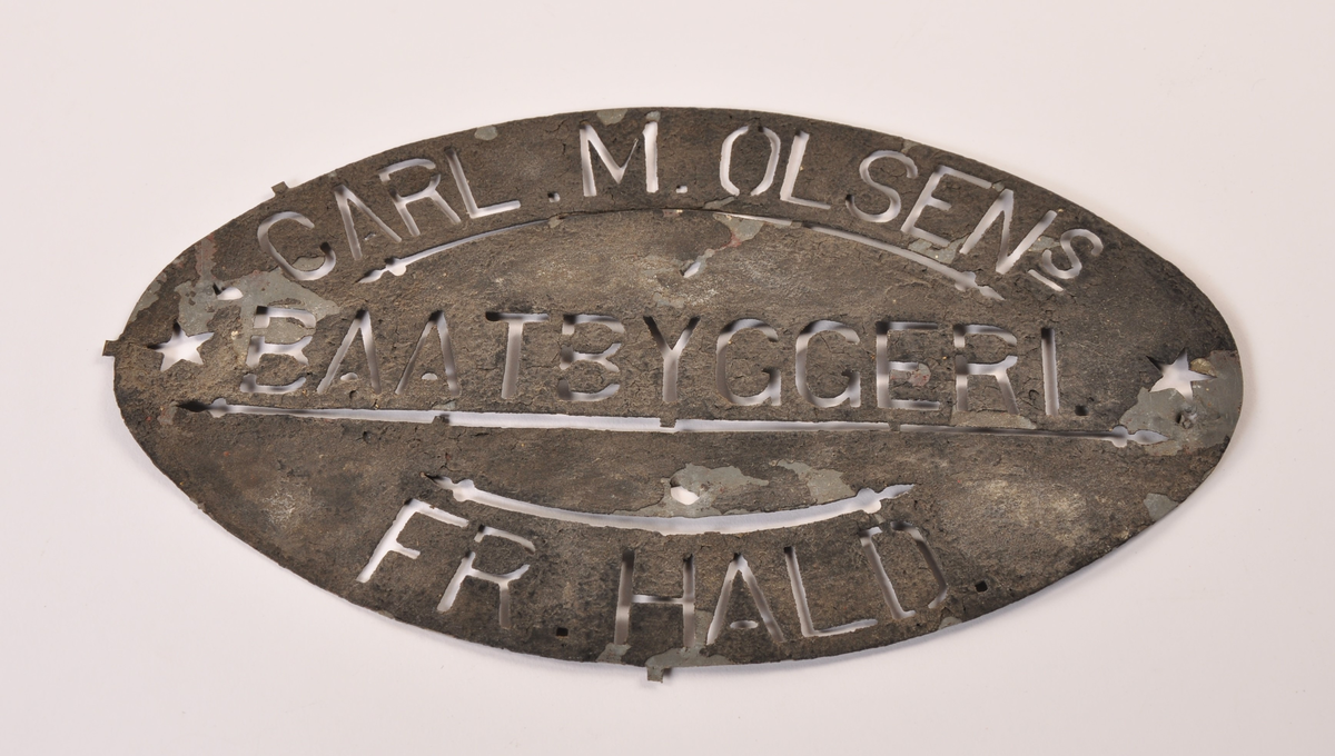 Oval metallsjablong med gjennombrudte bokstaver i metallet,  innskriften: Carl. M. Olsens Baatbyggeri. Fr. hald. Med løs, utstanset oval av rektangulær plate.
