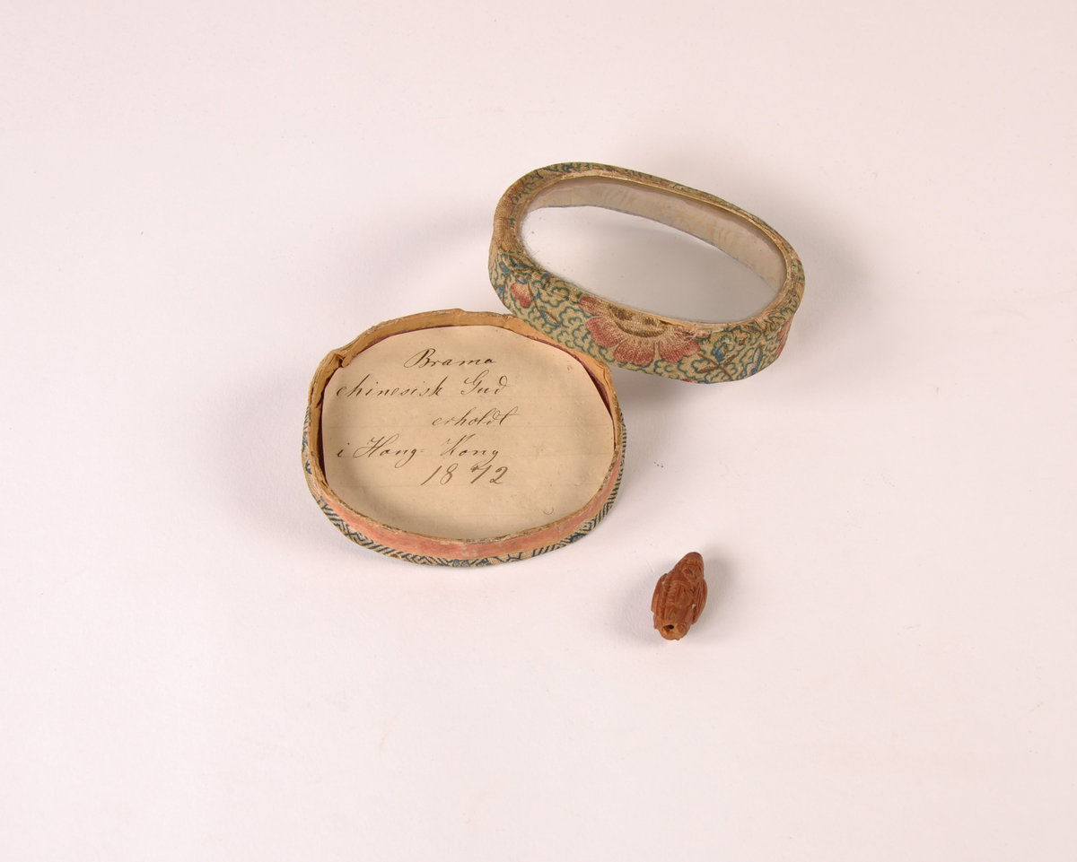 Oval boks treboks trukket med blomstret tekstil med glassplate. Inneholder en utskåret figur i rødt matriale.