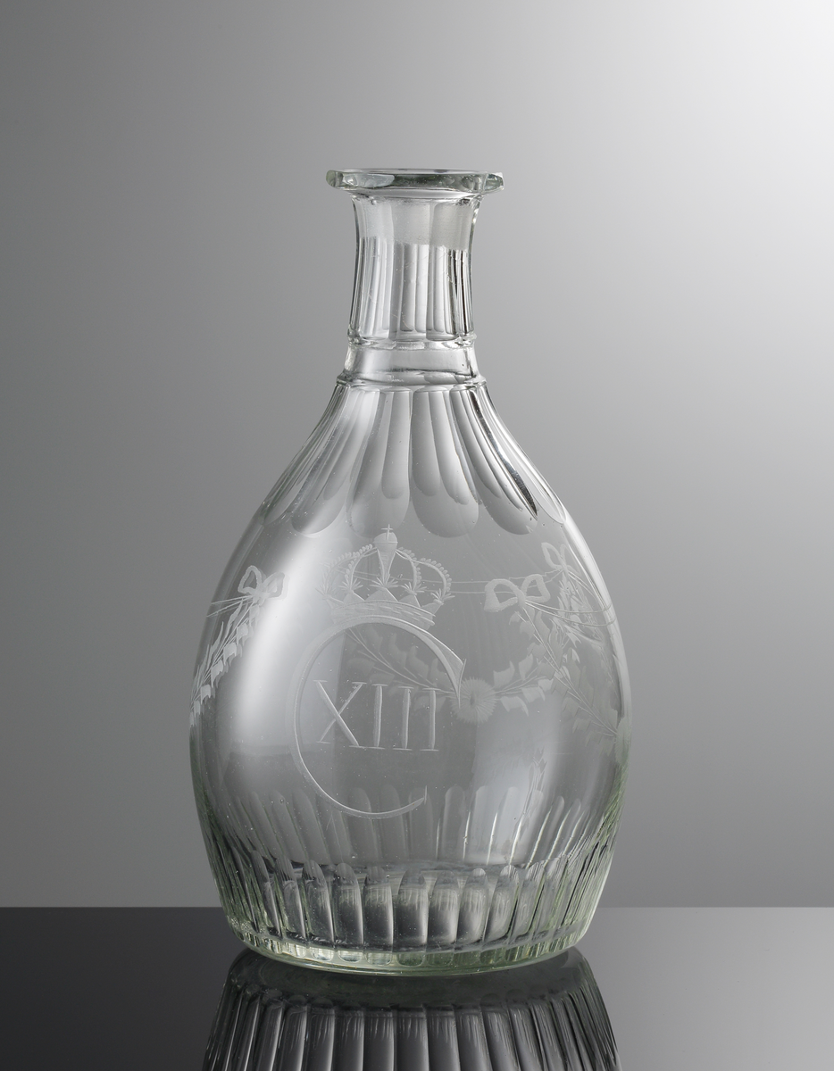 Karaff utan propp, tillhör "Carl XIII:s servis".
Blåst 1810-1815 ?. Graverad 1815-1818 ? (Carl XIII 1809 - 1818).
Graverad av Anders Spolander. Se E. Steenberg "Svenskt adertonhundratals glas", sidan 131, 1).
Bukig, tämligen kort hals, stor regalin. Regalin, hals och överdel är facettslipade. Nedsänkt ring runt halsen. Monogram "C XIII" med krona samt tre girlander i mittpartiet. En rad ställda oliver nedtill.
Ofärgat klarglas (dålig färg).
Blåst. Stor kula. Slipad och graverad dekor.

Inskrivet i huvudkatalogen tidigast 1976.

Enligt uppgift ägdes en del av servisen av Växjö regemente tills regementet upplöstes år 18??. Skänktes sedan till regementets officerare. Dessa pjäser tillhörde fänrik ....

Gravör Anders Spolander, född i Umeå den 27 juli 1782, arbetade som glassliparlärling vid Strömbäcks glasbruk 1806-1812, vid Kungsholms glasbruk 1812-1813, och 1813 vid en egen verkstad för "glasritning". Dog 1836. Hans verkstad drevs av änkan till 1843.
Funktion: Karaff