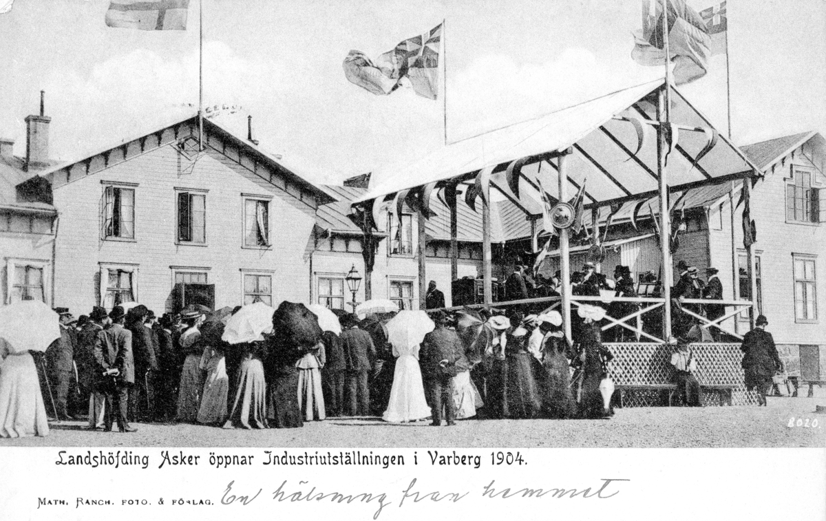 Brevkort, "Landshöfding Asker öppnar Industriutställningen i Varberg 1904" Folksamling på Gamlebyskolans gård nedanför en takförsedd scen, prydd med vajande unionsflaggor, där landshövdingen och en orkester står. Axel Asker var Hallands landshövding 1902–1916. Hantverks- och Industriutställningen 1904.