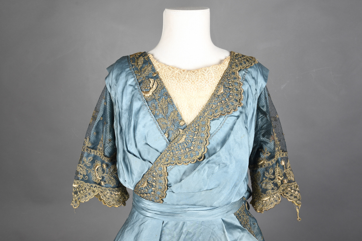 Fotsid kjole i lyseblå silke-duchesse. Blylodd nederst i kanten. Den har slep som er festet i livet, drapert og med blylodd nede. Kjolen er avdelt i livet og har drapert belte i kjolestoffet som lukkes med hekter. Den har 32 cm lange isydde ermer. Utover ermene er det trukket mørkere blå tyll brodert med gullfarget metalltråd. Stoffet på skuldrene er trukket noe utover ermene. Kjolen har V-utringning foran og bak, kantet med samme stoffet som er trukket utenpå ermene. V-utringningene er dekket med tynn hvit bomull belagt med kniplingsbroderi. Livet er fóret med hvit lerret. Kjolen lukkes bak med hekter og trykknapper.
