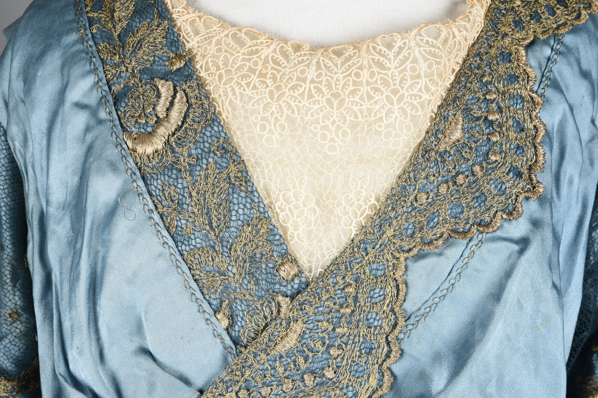 Fotsid kjole i lyseblå silke-duchesse. Blylodd nederst i kanten. Den har slep som er festet i livet, drapert og med blylodd nede. Kjolen er avdelt i livet og har drapert belte i kjolestoffet som lukkes med hekter. Den har 32 cm lange isydde ermer. Utover ermene er det trukket mørkere blå tyll brodert med gullfarget metalltråd. Stoffet på skuldrene er trukket noe utover ermene. Kjolen har V-utringning foran og bak, kantet med samme stoffet som er trukket utenpå ermene. V-utringningene er dekket med tynn hvit bomull belagt med kniplingsbroderi. Livet er fóret med hvit lerret. Kjolen lukkes bak med hekter og trykknapper.
