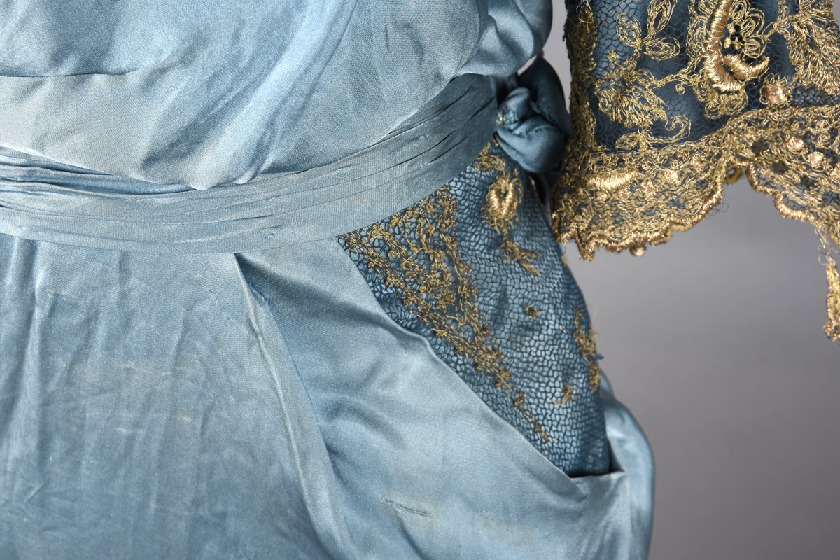 Fotsid kjole i lyseblå silke-duchesse. Blylodd nederst i kanten. Den har slep som er festet i livet, drapert og med blylodd nede. Kjolen er avdelt i livet og har drapert belte i kjolestoffet som lukkes med hekter. Den har 32 cm lange isydde ermer. Utover ermene er det trukket mørkere blå tyll brodert med gullfarget metalltråd. Stoffet på skuldrene er trukket noe utover ermene. Kjolen har V-utringning foran og bak, kantet med samme stoffet som er trukket utenpå ermene. V-utringningene er dekket med tynn hvit bomull belagt med kniplingsbroderi. Livet er fóret med hvit lerret. Kjolen lukkes bak med hekter og trykknapper.