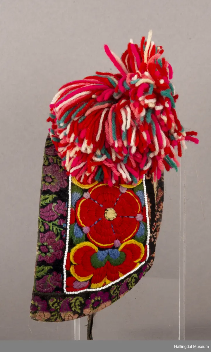Brodert på svart fløyel. Tett brodert med ullgarn, hallingsaum, roser i fargene raudt, 2 gulfarger, grønt blått og fiolett. Rundt etter kanten boler, fløyelsband med svart bottfarge og vove mønster i fargane grønt og mørk fiolett, blått og lys raud-orange. Bak pannelinet er det kanta med eit blomstrut fløyelstykke (svart, raudbrunt og lys orange). Mellom  broderinga og bolene er det ei rad med perler - kårilsteiner. 2 band 12,5 cm lange, til å knyte fast pannelinet med. Ubleika bomullstoff til for. Panneskuve (dusk) av flanillgarn, fargear i skuven: kvitt, gult, orange, rosa og grønt.