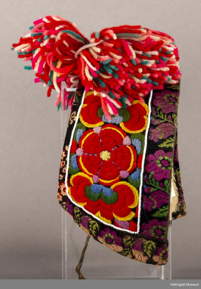 Brodert på svart fløyel. Tett brodert med ullgarn, hallingsaum, roser i fargene raudt, 2 gulfarger, grønt blått og fiolett. Rundt etter kanten boler, fløyelsband med svart bottfarge og vove mønster i fargane grønt og mørk fiolett, blått og lys raud-orange. Bak pannelinet er det kanta med eit blomstrut fløyelstykke (svart, raudbrunt og lys orange). Mellom  broderinga og bolene er det ei rad med perler - kårilsteiner. 2 band 12,5 cm lange, til å knyte fast pannelinet med. Ubleika bomullstoff til for. Panneskuve (dusk) av flanillgarn, fargear i skuven: kvitt, gult, orange, rosa og grønt.