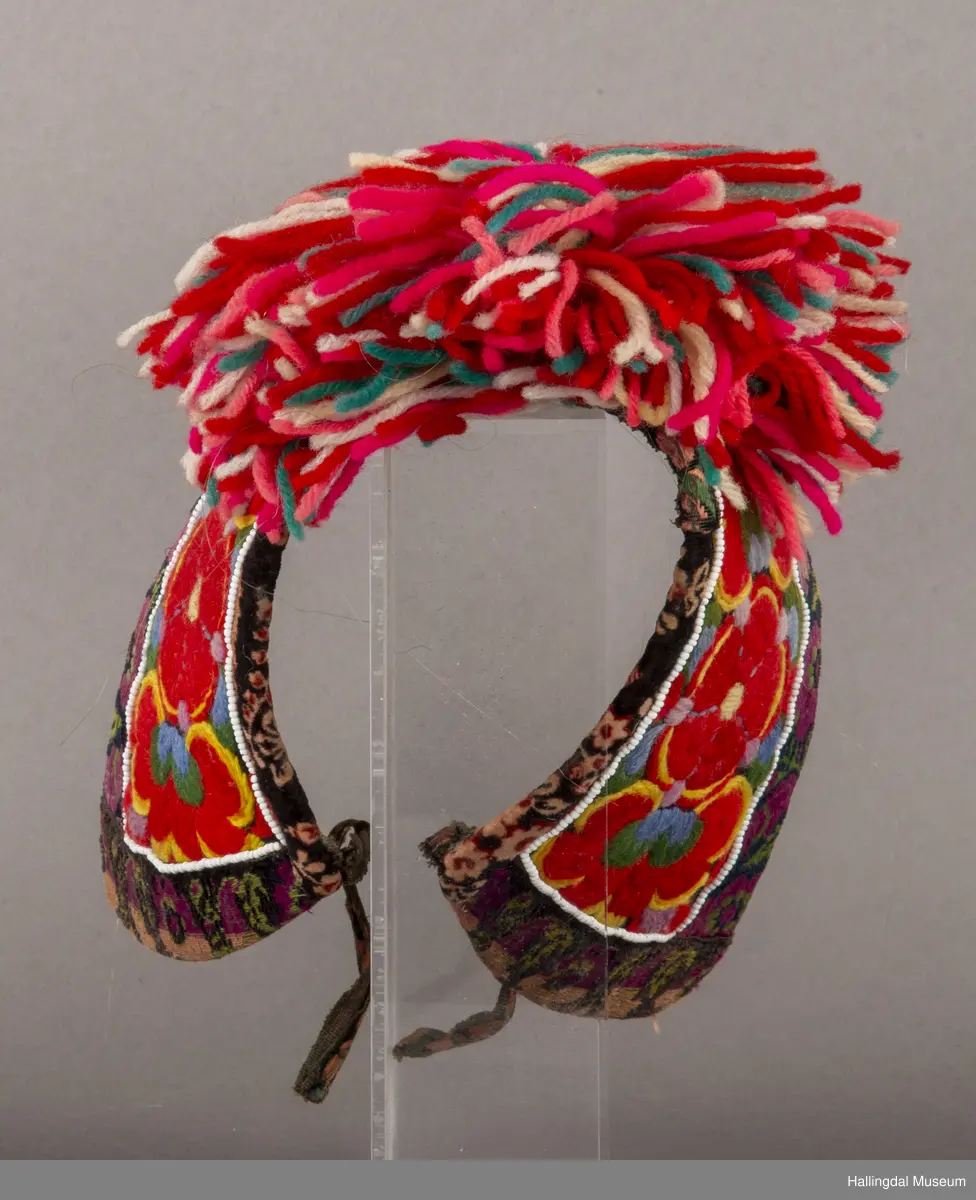 Brodert på svart fløyel. Tett brodert med ullgarn, hallingsaum, roser i fargene raudt, 2 gulfarger, grønt blått og fiolett. Rundt etter kanten boler, fløyelsband med svart bottfarge og vove mønster i fargane grønt og mørk fiolett, blått og lys raud-orange. Bak pannelinet er det kanta med eit blomstrut fløyelstykke (svart, raudbrunt og lys orange). Mellom  broderinga og bolene er det ei rad med perler - kårilsteiner. 2 band 12,5 cm lange, til å knyte fast pannelinet med. Ubleika bomullstoff til for. Panneskuve (dusk) av flanillgarn, fargear i skuven: kvitt, gult, orange, rosa og grønt.