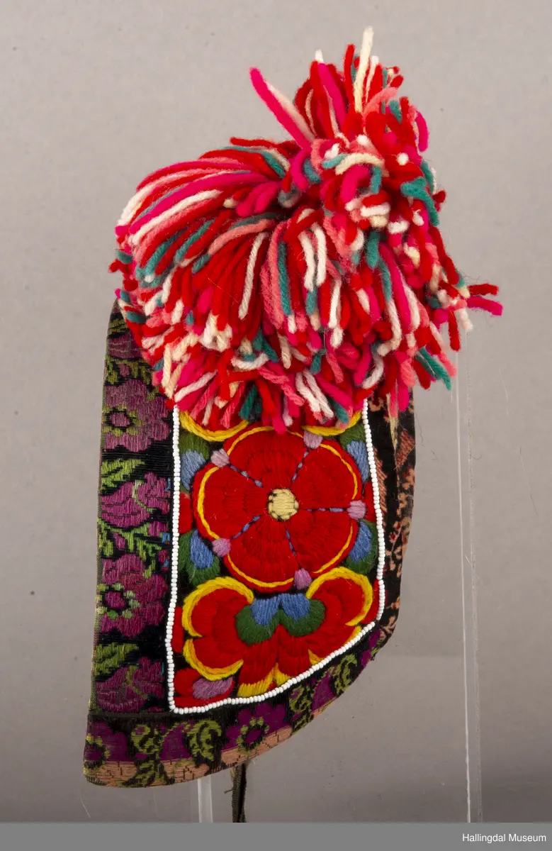 Brodert på svart fløyel. Tett brodert med ullgarn, hallingsaum, roser i fargene raudt, 2 gulfarger, grønt blått og fiolett. Rundt etter kanten boler, fløyelsband med svart bottfarge og vove mønster i fargane grønt og mørk fiolett, blått og lys raud-orange. Bak pannelinet er det kanta med eit blomstrut fløyelstykke (svart, raudbrunt og lys orange). Mellom  broderinga og bolene er det ei rad med perler - kårilsteiner. 2 band 12,5 cm lange, til å knyte fast pannelinet med. Ubleika bomullstoff til for. Panneskuve (dusk) av flanillgarn, fargear i skuven: kvitt, gult, orange, rosa og grønt.