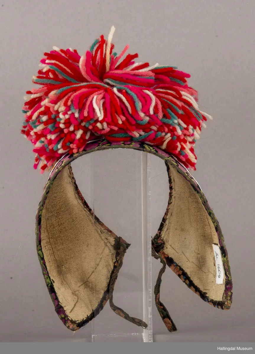 Brodert på svart fløyel. Tett brodert med ullgarn, hallingsaum, roser i fargene raudt, 2 gulfarger, grønt blått og fiolett. Rundt etter kanten boler, fløyelsband med svart bottfarge og vove mønster i fargane grønt og mørk fiolett, blått og lys raud-orange. Bak pannelinet er det kanta med eit blomstrut fløyelstykke (svart, raudbrunt og lys orange). Mellom  broderinga og bolene er det ei rad med perler - kårilsteiner. 2 band 12,5 cm lange, til å knyte fast pannelinet med. Ubleika bomullstoff til for. Panneskuve (dusk) av flanillgarn, fargear i skuven: kvitt, gult, orange, rosa og grønt.