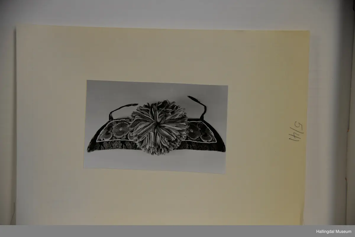 Brodert på svart fløyel. Tett brodert med ullgarn, hallingsaum, roser i fargene raudt, 2 gulfarger, grønt blått og fiolett. Rundt etter kanten boler, fløyelsband med svart bottfarge og vove mønster i fargane grønt og mørk fiolett, blått og lys raud-orange. Bak pannelinet er det kanta med eit blomstrut fløyelstykke (svart, raudbrunt og lys orange). Mellom  broderinga og bolene er det ei rad med perler - kårilsteiner. 2 band 12,5 cm lange, til å knyte fast pannelinet med. Ubleika bomullstoff til for. Panneskuve (dusk) av flanillgarn, fargear i skuven: kvitt, gult, orange, rosa og grønt.