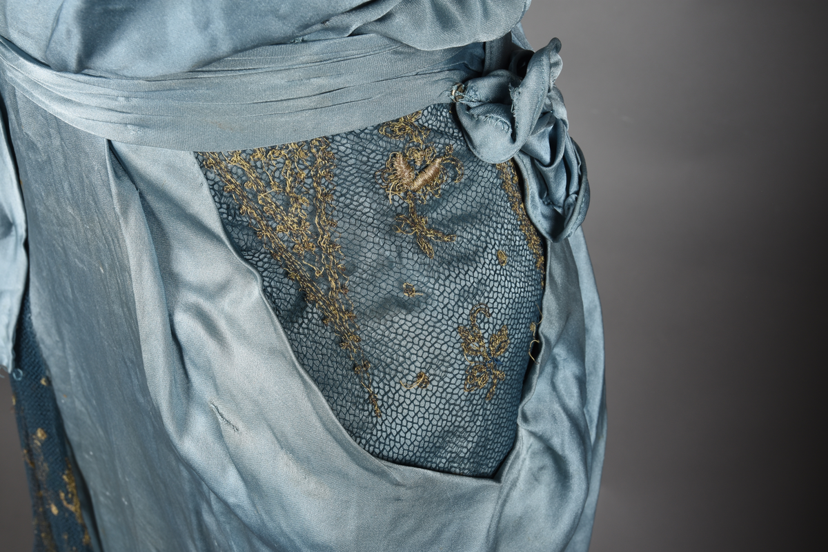 Fotsid kjole i lyseblå silke-duchesse. Blylodd nederst i kanten. Den har slep som er festet i livet, drapert og med blylodd nede. Kjolen er avdelt i livet og har drapert belte i kjolestoffet som lukkes med hekter. Den har 32 cm lange isydde ermer. Utover ermene er det trukket mørkere blå tyll brodert med gullfarget metalltråd. Stoffet på skuldrene er trukket noe utover ermene. Kjolen har V-utringning foran og bak, kantet med samme stoffet som er trukket utenpå ermene. V-utringningene er dekket med tynn hvit bomull belagt med kniplingsbroderi. Livet er fóret med hvit lerret. Kjolen lukkes bak med hekter og trykknapper.