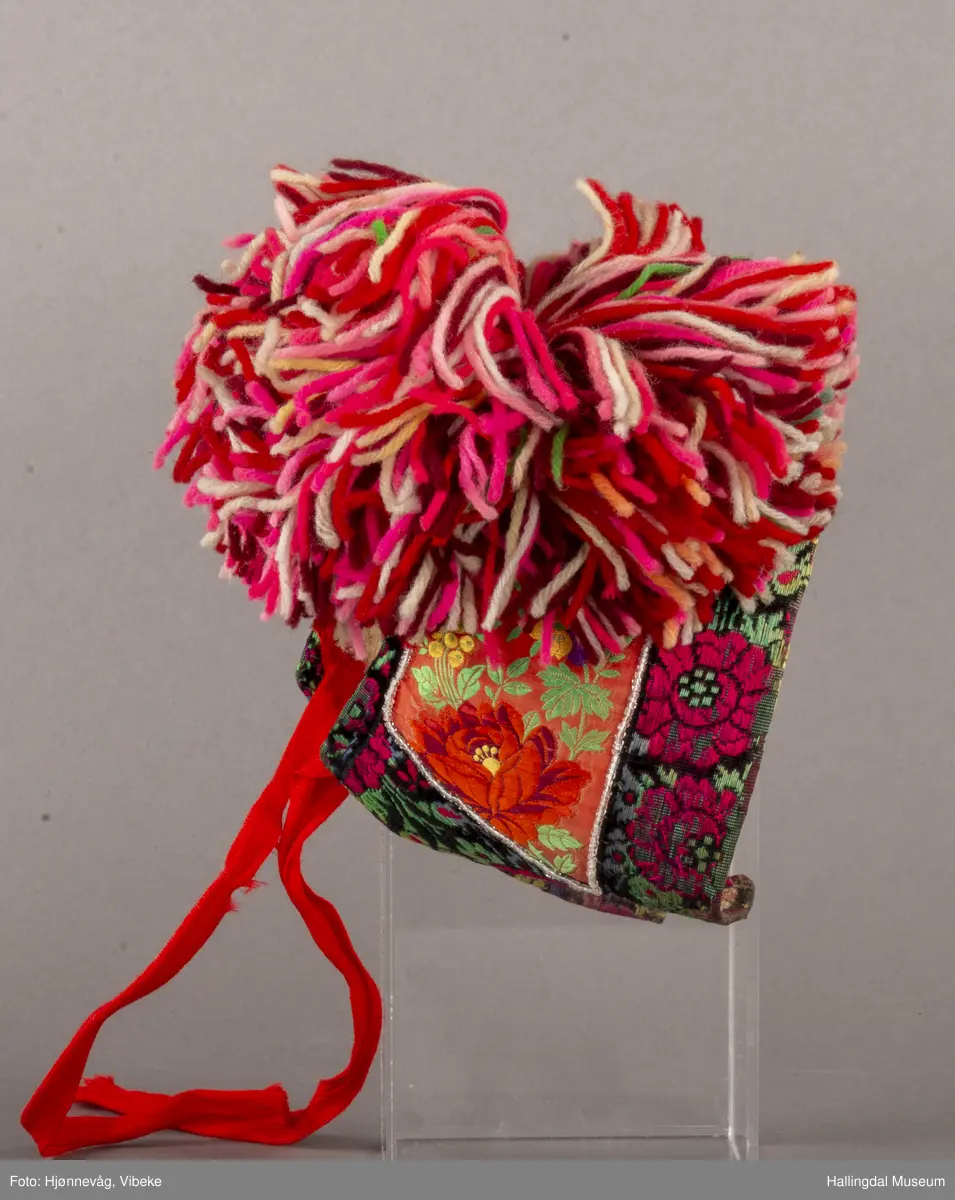 a) Jentepannelin av papp, sylkjety, bolu, kårilsteinar, bomullsty, ullgarn, sydd.
b) Nakkeskuve av flanillsgarn i raudt, mørk raudt, lys raudt, kvitt, lys gult, og grønt.