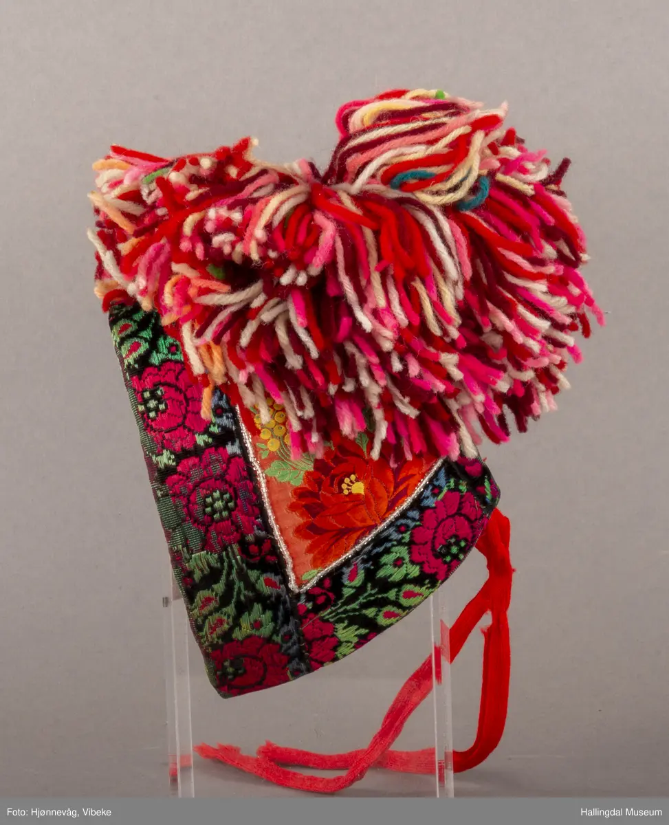 a) Jentepannelin av papp, sylkjety, bolu, kårilsteinar, bomullsty, ullgarn, sydd.
b) Nakkeskuve av flanillsgarn i raudt, mørk raudt, lys raudt, kvitt, lys gult, og grønt.