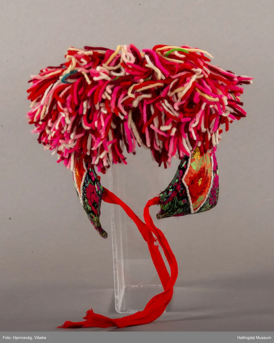 a) Jentepannelin av papp, sylkjety, bolu, kårilsteinar, bomullsty, ullgarn, sydd.
b) Nakkeskuve av flanillsgarn i raudt, mørk raudt, lys raudt, kvitt, lys gult, og grønt.