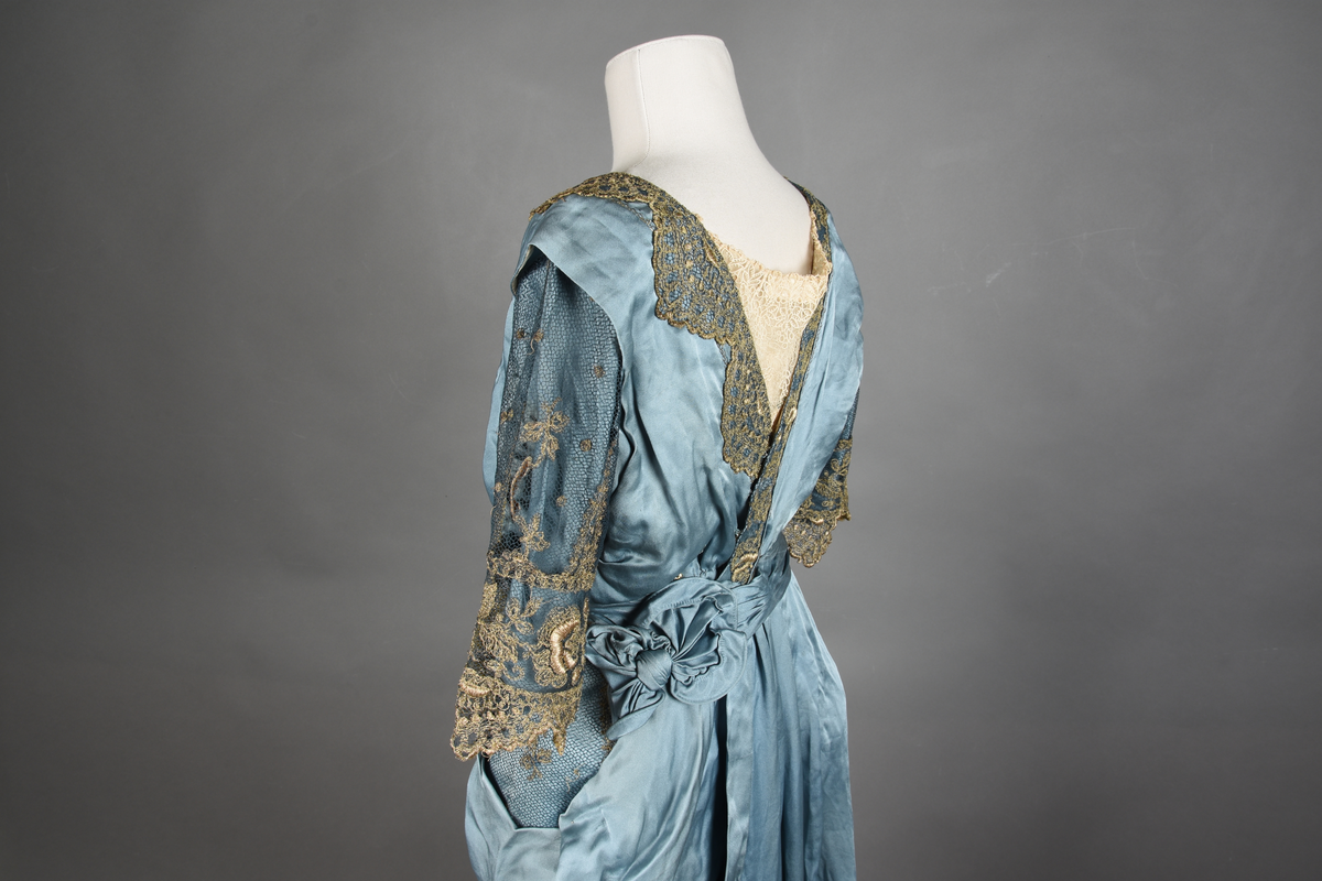 Fotsid kjole i lyseblå silke-duchesse. Blylodd nederst i kanten. Den har slep som er festet i livet, drapert og med blylodd nede. Kjolen er avdelt i livet og har drapert belte i kjolestoffet som lukkes med hekter. Den har 32 cm lange isydde ermer. Utover ermene er det trukket mørkere blå tyll brodert med gullfarget metalltråd. Stoffet på skuldrene er trukket noe utover ermene. Kjolen har V-utringning foran og bak, kantet med samme stoffet som er trukket utenpå ermene. V-utringningene er dekket med tynn hvit bomull belagt med kniplingsbroderi. Livet er fóret med hvit lerret. Kjolen lukkes bak med hekter og trykknapper.