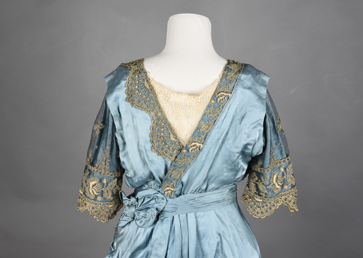 Fotsid kjole i lyseblå silke-duchesse. Blylodd nederst i kanten. Den har slep som er festet i livet, drapert og med blylodd nede. Kjolen er avdelt i livet og har drapert belte i kjolestoffet som lukkes med hekter. Den har 32 cm lange isydde ermer. Utover ermene er det trukket mørkere blå tyll brodert med gullfarget metalltråd. Stoffet på skuldrene er trukket noe utover ermene. Kjolen har V-utringning foran og bak, kantet med samme stoffet som er trukket utenpå ermene. V-utringningene er dekket med tynn hvit bomull belagt med kniplingsbroderi. Livet er fóret med hvit lerret. Kjolen lukkes bak med hekter og trykknapper.