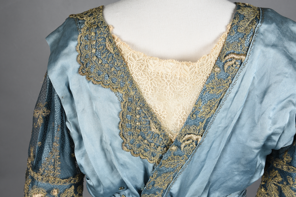 Fotsid kjole i lyseblå silke-duchesse. Blylodd nederst i kanten. Den har slep som er festet i livet, drapert og med blylodd nede. Kjolen er avdelt i livet og har drapert belte i kjolestoffet som lukkes med hekter. Den har 32 cm lange isydde ermer. Utover ermene er det trukket mørkere blå tyll brodert med gullfarget metalltråd. Stoffet på skuldrene er trukket noe utover ermene. Kjolen har V-utringning foran og bak, kantet med samme stoffet som er trukket utenpå ermene. V-utringningene er dekket med tynn hvit bomull belagt med kniplingsbroderi. Livet er fóret med hvit lerret. Kjolen lukkes bak med hekter og trykknapper.
