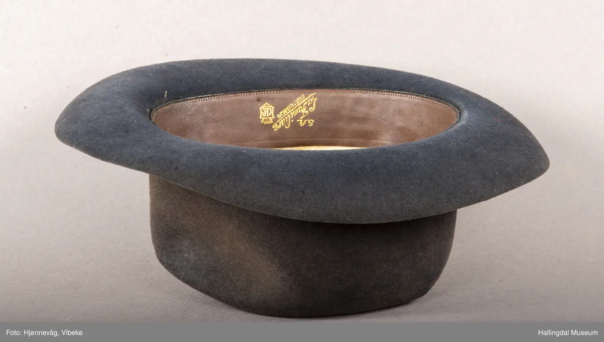 Filthatt med et bredt silkebånd rundt pullen. Høyde: 12 cm, Bredde brem: 6,5 cm. Gråbrun.
