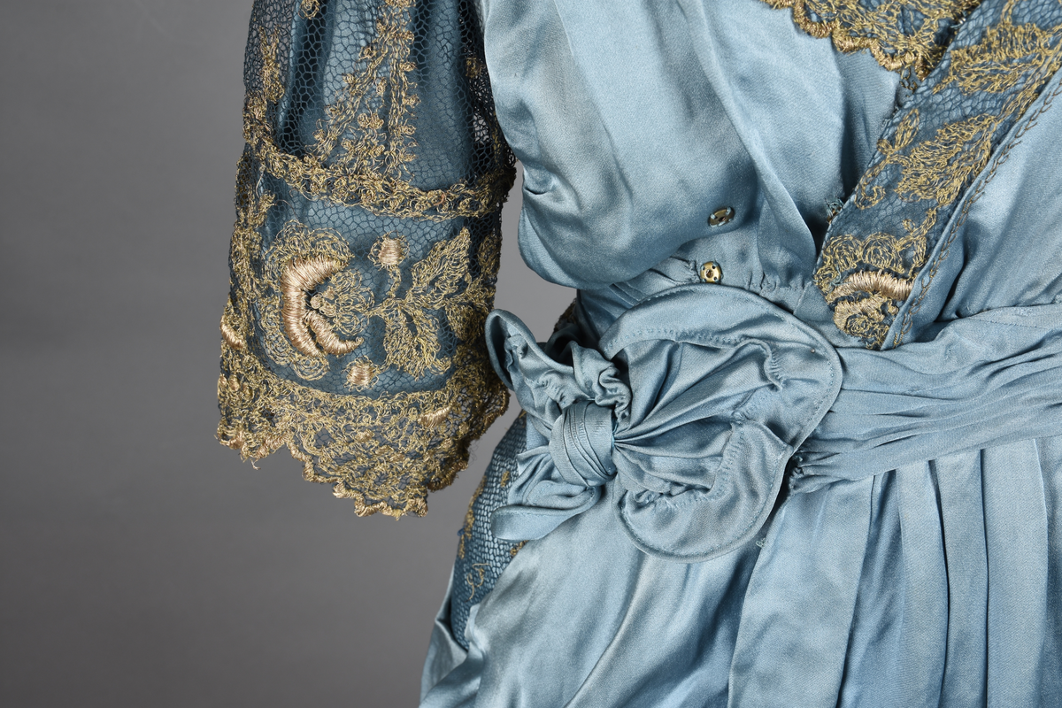 Fotsid kjole i lyseblå silke-duchesse. Blylodd nederst i kanten. Den har slep som er festet i livet, drapert og med blylodd nede. Kjolen er avdelt i livet og har drapert belte i kjolestoffet som lukkes med hekter. Den har 32 cm lange isydde ermer. Utover ermene er det trukket mørkere blå tyll brodert med gullfarget metalltråd. Stoffet på skuldrene er trukket noe utover ermene. Kjolen har V-utringning foran og bak, kantet med samme stoffet som er trukket utenpå ermene. V-utringningene er dekket med tynn hvit bomull belagt med kniplingsbroderi. Livet er fóret med hvit lerret. Kjolen lukkes bak med hekter og trykknapper.