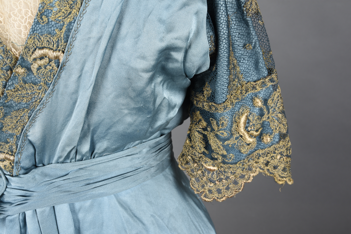 Fotsid kjole i lyseblå silke-duchesse. Blylodd nederst i kanten. Den har slep som er festet i livet, drapert og med blylodd nede. Kjolen er avdelt i livet og har drapert belte i kjolestoffet som lukkes med hekter. Den har 32 cm lange isydde ermer. Utover ermene er det trukket mørkere blå tyll brodert med gullfarget metalltråd. Stoffet på skuldrene er trukket noe utover ermene. Kjolen har V-utringning foran og bak, kantet med samme stoffet som er trukket utenpå ermene. V-utringningene er dekket med tynn hvit bomull belagt med kniplingsbroderi. Livet er fóret med hvit lerret. Kjolen lukkes bak med hekter og trykknapper.