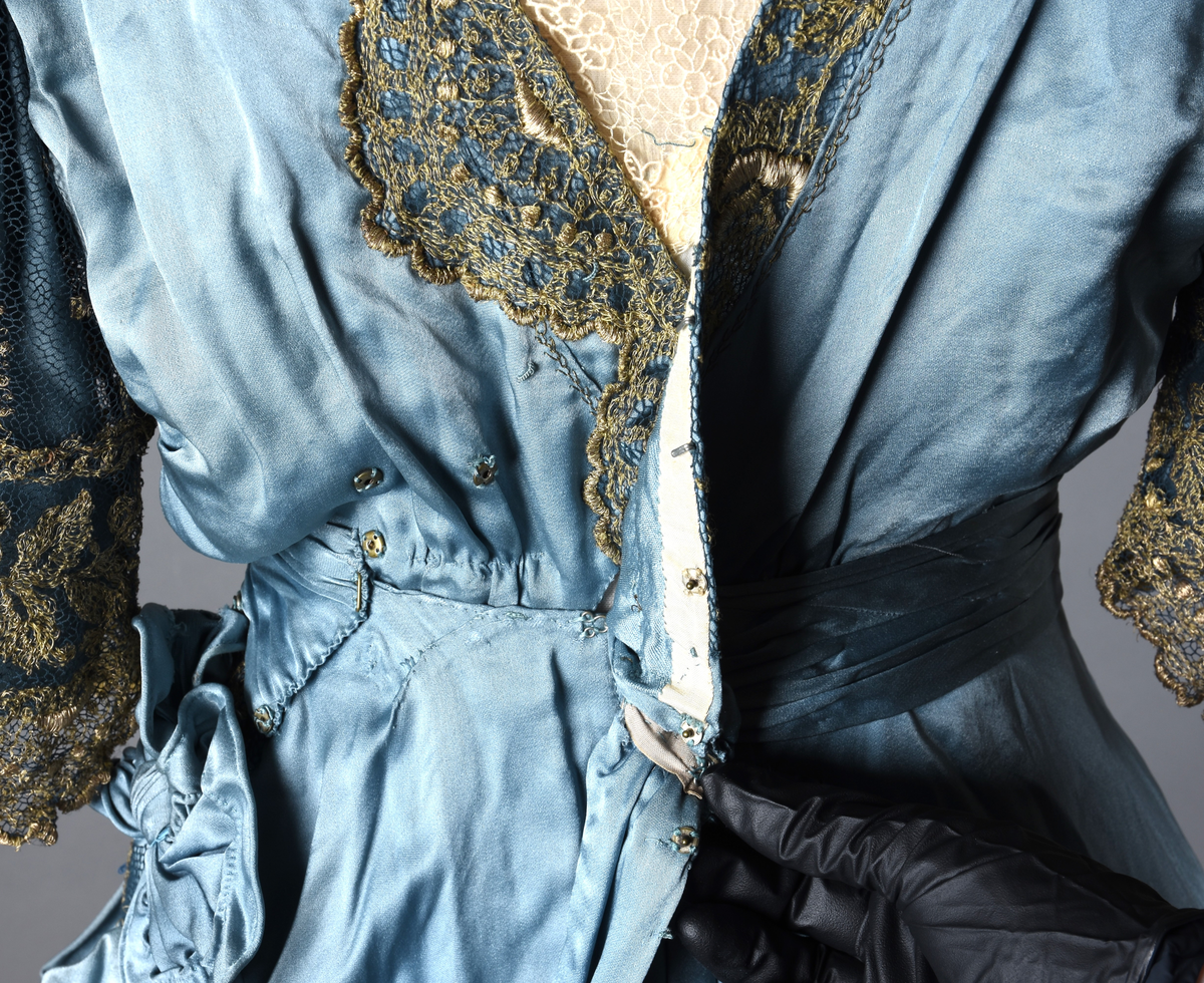 Fotsid kjole i lyseblå silke-duchesse. Blylodd nederst i kanten. Den har slep som er festet i livet, drapert og med blylodd nede. Kjolen er avdelt i livet og har drapert belte i kjolestoffet som lukkes med hekter. Den har 32 cm lange isydde ermer. Utover ermene er det trukket mørkere blå tyll brodert med gullfarget metalltråd. Stoffet på skuldrene er trukket noe utover ermene. Kjolen har V-utringning foran og bak, kantet med samme stoffet som er trukket utenpå ermene. V-utringningene er dekket med tynn hvit bomull belagt med kniplingsbroderi. Livet er fóret med hvit lerret. Kjolen lukkes bak med hekter og trykknapper.