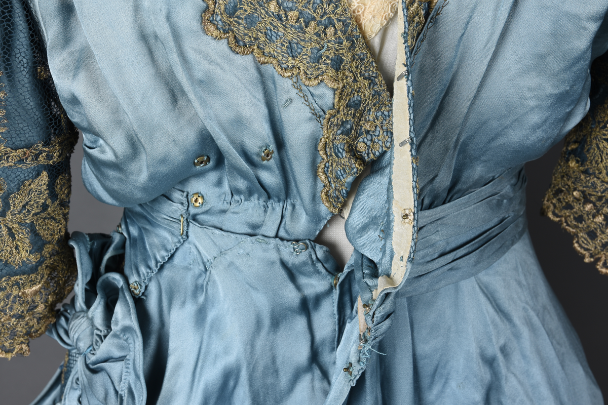Fotsid kjole i lyseblå silke-duchesse. Blylodd nederst i kanten. Den har slep som er festet i livet, drapert og med blylodd nede. Kjolen er avdelt i livet og har drapert belte i kjolestoffet som lukkes med hekter. Den har 32 cm lange isydde ermer. Utover ermene er det trukket mørkere blå tyll brodert med gullfarget metalltråd. Stoffet på skuldrene er trukket noe utover ermene. Kjolen har V-utringning foran og bak, kantet med samme stoffet som er trukket utenpå ermene. V-utringningene er dekket med tynn hvit bomull belagt med kniplingsbroderi. Livet er fóret med hvit lerret. Kjolen lukkes bak med hekter og trykknapper.