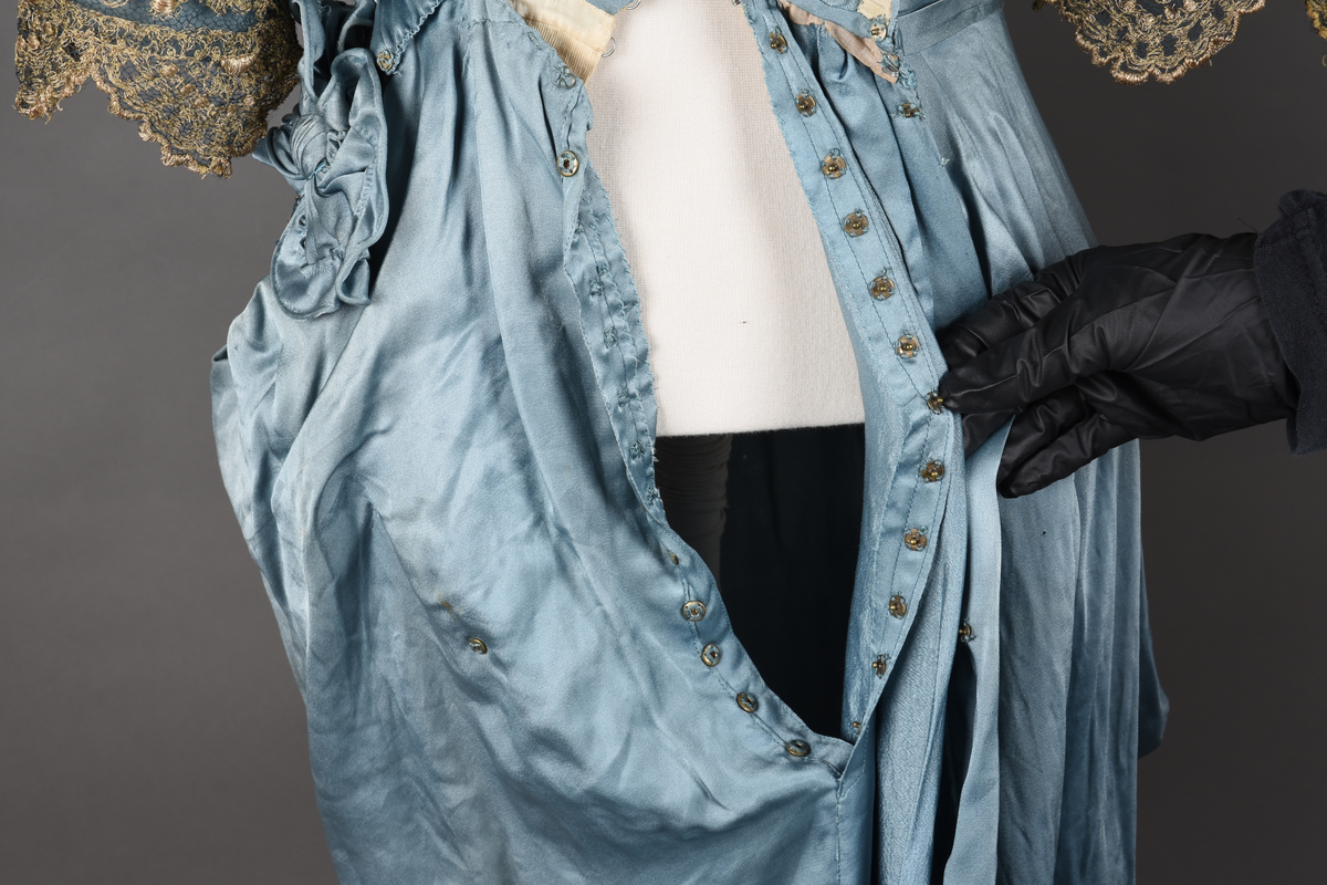 Fotsid kjole i lyseblå silke-duchesse. Blylodd nederst i kanten. Den har slep som er festet i livet, drapert og med blylodd nede. Kjolen er avdelt i livet og har drapert belte i kjolestoffet som lukkes med hekter. Den har 32 cm lange isydde ermer. Utover ermene er det trukket mørkere blå tyll brodert med gullfarget metalltråd. Stoffet på skuldrene er trukket noe utover ermene. Kjolen har V-utringning foran og bak, kantet med samme stoffet som er trukket utenpå ermene. V-utringningene er dekket med tynn hvit bomull belagt med kniplingsbroderi. Livet er fóret med hvit lerret. Kjolen lukkes bak med hekter og trykknapper.