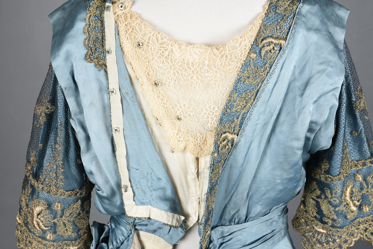 Fotsid kjole i lyseblå silke-duchesse. Blylodd nederst i kanten. Den har slep som er festet i livet, drapert og med blylodd nede. Kjolen er avdelt i livet og har drapert belte i kjolestoffet som lukkes med hekter. Den har 32 cm lange isydde ermer. Utover ermene er det trukket mørkere blå tyll brodert med gullfarget metalltråd. Stoffet på skuldrene er trukket noe utover ermene. Kjolen har V-utringning foran og bak, kantet med samme stoffet som er trukket utenpå ermene. V-utringningene er dekket med tynn hvit bomull belagt med kniplingsbroderi. Livet er fóret med hvit lerret. Kjolen lukkes bak med hekter og trykknapper.