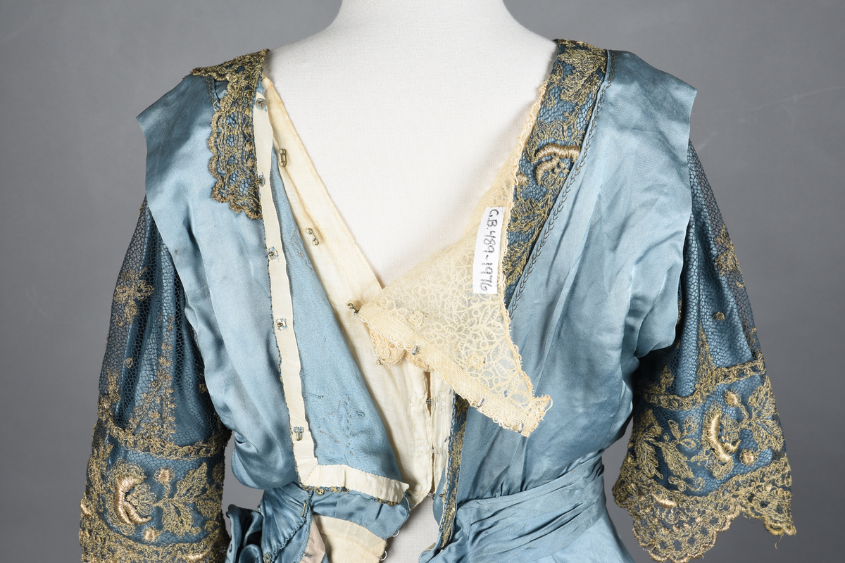 Fotsid kjole i lyseblå silke-duchesse. Blylodd nederst i kanten. Den har slep som er festet i livet, drapert og med blylodd nede. Kjolen er avdelt i livet og har drapert belte i kjolestoffet som lukkes med hekter. Den har 32 cm lange isydde ermer. Utover ermene er det trukket mørkere blå tyll brodert med gullfarget metalltråd. Stoffet på skuldrene er trukket noe utover ermene. Kjolen har V-utringning foran og bak, kantet med samme stoffet som er trukket utenpå ermene. V-utringningene er dekket med tynn hvit bomull belagt med kniplingsbroderi. Livet er fóret med hvit lerret. Kjolen lukkes bak med hekter og trykknapper.