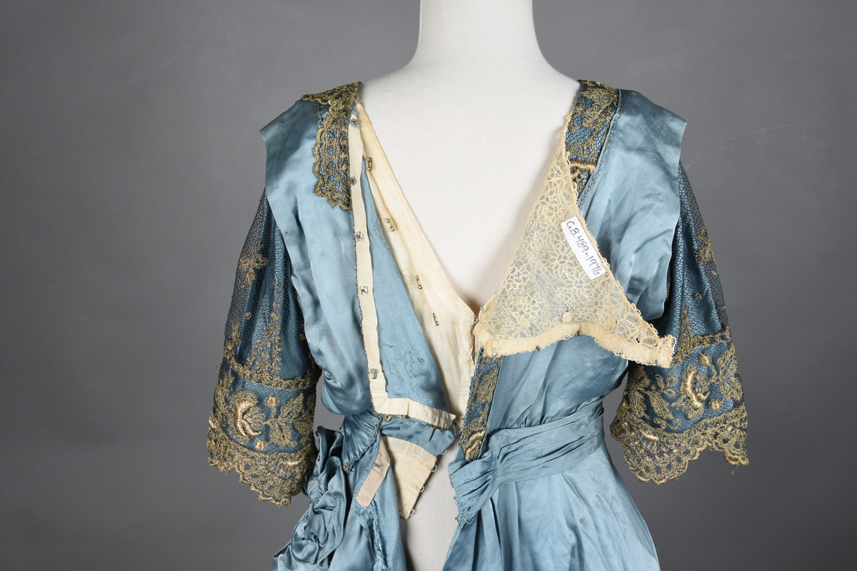 Fotsid kjole i lyseblå silke-duchesse. Blylodd nederst i kanten. Den har slep som er festet i livet, drapert og med blylodd nede. Kjolen er avdelt i livet og har drapert belte i kjolestoffet som lukkes med hekter. Den har 32 cm lange isydde ermer. Utover ermene er det trukket mørkere blå tyll brodert med gullfarget metalltråd. Stoffet på skuldrene er trukket noe utover ermene. Kjolen har V-utringning foran og bak, kantet med samme stoffet som er trukket utenpå ermene. V-utringningene er dekket med tynn hvit bomull belagt med kniplingsbroderi. Livet er fóret med hvit lerret. Kjolen lukkes bak med hekter og trykknapper.