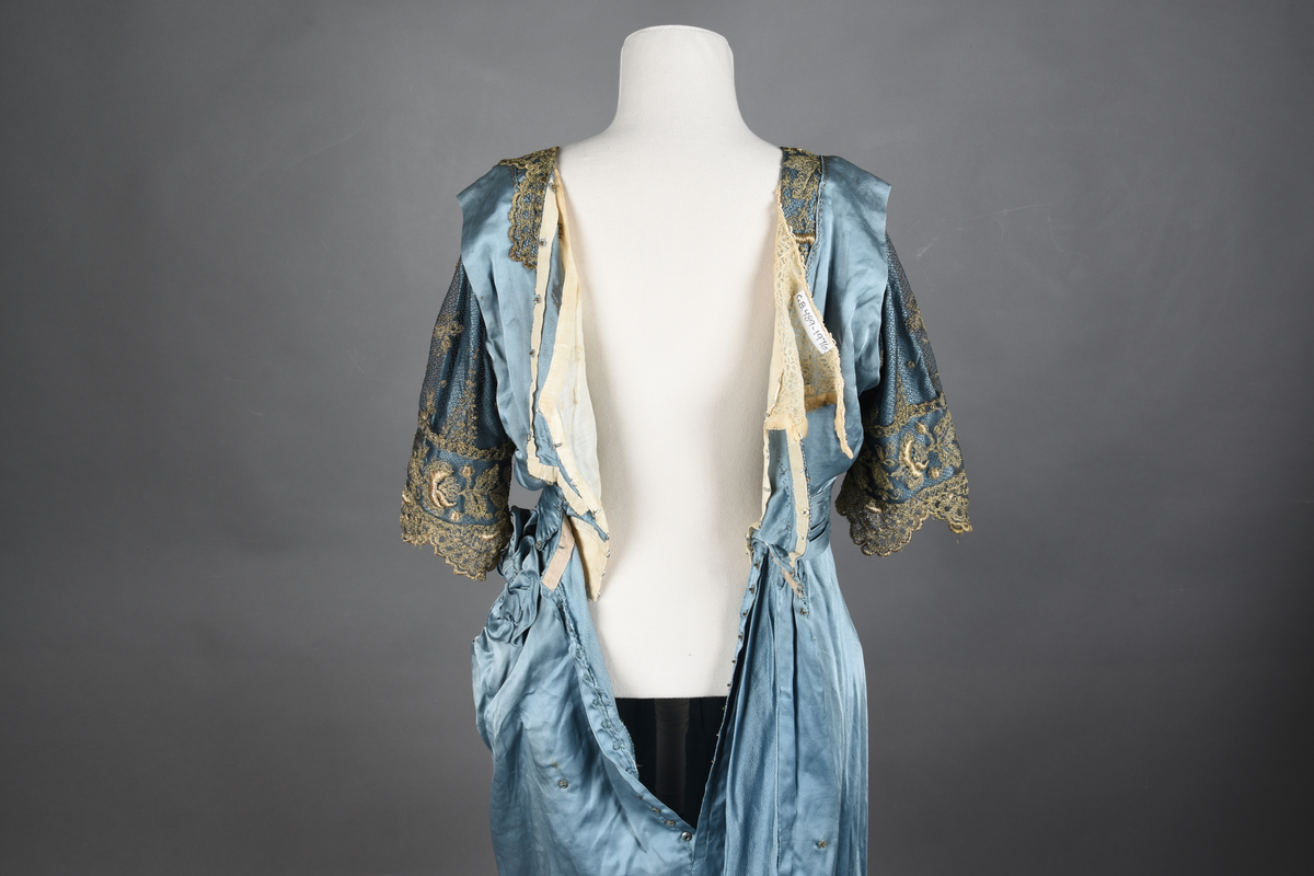 Fotsid kjole i lyseblå silke-duchesse. Blylodd nederst i kanten. Den har slep som er festet i livet, drapert og med blylodd nede. Kjolen er avdelt i livet og har drapert belte i kjolestoffet som lukkes med hekter. Den har 32 cm lange isydde ermer. Utover ermene er det trukket mørkere blå tyll brodert med gullfarget metalltråd. Stoffet på skuldrene er trukket noe utover ermene. Kjolen har V-utringning foran og bak, kantet med samme stoffet som er trukket utenpå ermene. V-utringningene er dekket med tynn hvit bomull belagt med kniplingsbroderi. Livet er fóret med hvit lerret. Kjolen lukkes bak med hekter og trykknapper.