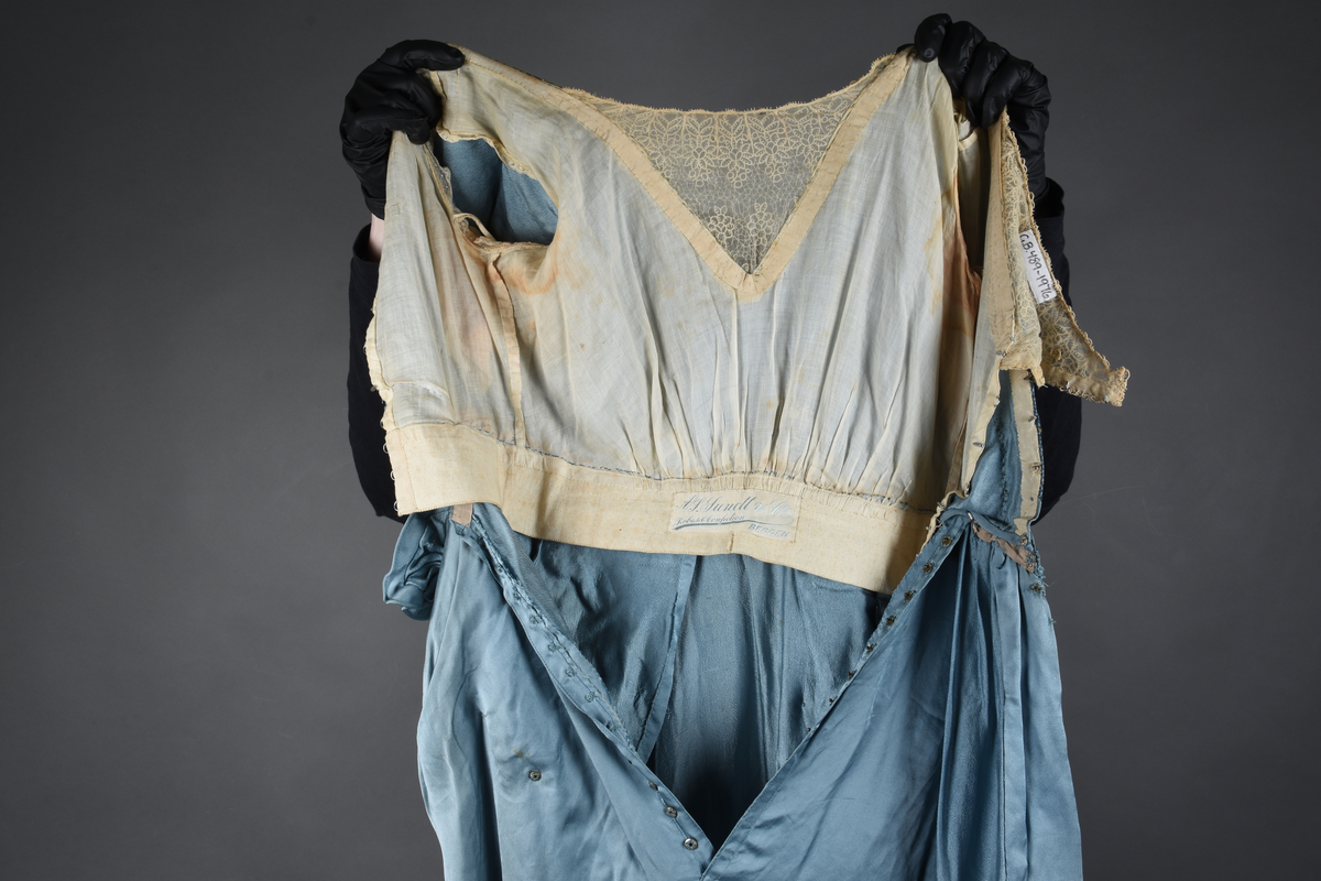 Fotsid kjole i lyseblå silke-duchesse. Blylodd nederst i kanten. Den har slep som er festet i livet, drapert og med blylodd nede. Kjolen er avdelt i livet og har drapert belte i kjolestoffet som lukkes med hekter. Den har 32 cm lange isydde ermer. Utover ermene er det trukket mørkere blå tyll brodert med gullfarget metalltråd. Stoffet på skuldrene er trukket noe utover ermene. Kjolen har V-utringning foran og bak, kantet med samme stoffet som er trukket utenpå ermene. V-utringningene er dekket med tynn hvit bomull belagt med kniplingsbroderi. Livet er fóret med hvit lerret. Kjolen lukkes bak med hekter og trykknapper.
