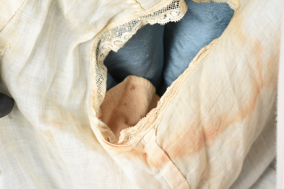 Fotsid kjole i lyseblå silke-duchesse. Blylodd nederst i kanten. Den har slep som er festet i livet, drapert og med blylodd nede. Kjolen er avdelt i livet og har drapert belte i kjolestoffet som lukkes med hekter. Den har 32 cm lange isydde ermer. Utover ermene er det trukket mørkere blå tyll brodert med gullfarget metalltråd. Stoffet på skuldrene er trukket noe utover ermene. Kjolen har V-utringning foran og bak, kantet med samme stoffet som er trukket utenpå ermene. V-utringningene er dekket med tynn hvit bomull belagt med kniplingsbroderi. Livet er fóret med hvit lerret. Kjolen lukkes bak med hekter og trykknapper.