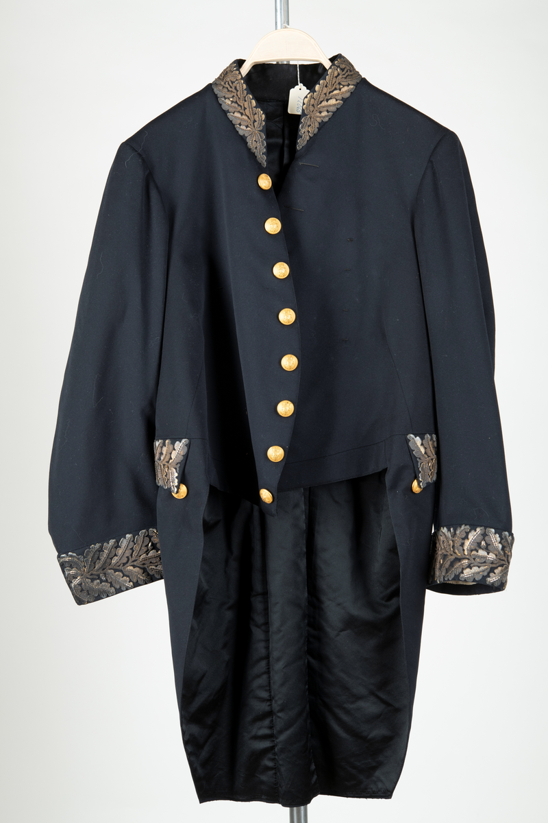 Paraduniformsrock till landshövdingeuniform. Mörkblå frackrock, v-ringad med ståkrage. Kragen, ärmslag och ficklock har broderade eklöv i gulmetalltråd. Knäppt fram med en hyska och hake. Åtta stycken garneringsknappar med motsvarande markerade knapphål. Knappar i gylmetall med krönt örn i hertigkrona. Livsömmar och skörtveck bak. Två knappar samt två ficklock med vardera tre knappar i midjesömmen bak. Tvåsömsärm med blindslag. Vadderad, foder av svart atlassiden. Fickor i skörtets foder.